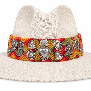 May include: A cream-colored straw hat with a wide brim. The hat features a colorful woven band with silver heart and flame charms. The band is a mix of red, yellow, and green. The hat is a stylish accessory.