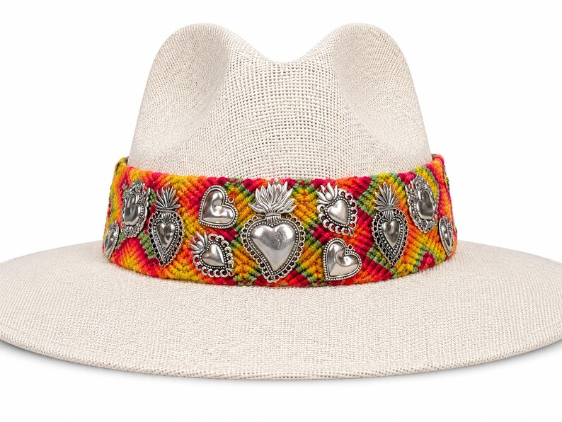 May include: A cream-colored straw hat with a wide brim. The hat features a colorful woven band with silver heart and flame charms. The band is a mix of red, yellow, and green. The hat is a stylish accessory.