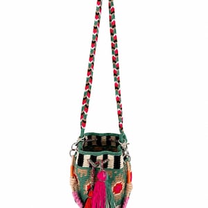 May include: A bucket bag featuring a woven green, black, and beige geometric pattern with a long braided strap in pink, green, white, and black. The bag includes a drawstring closure with multiple hanging tassels in red, pink, and green colors.