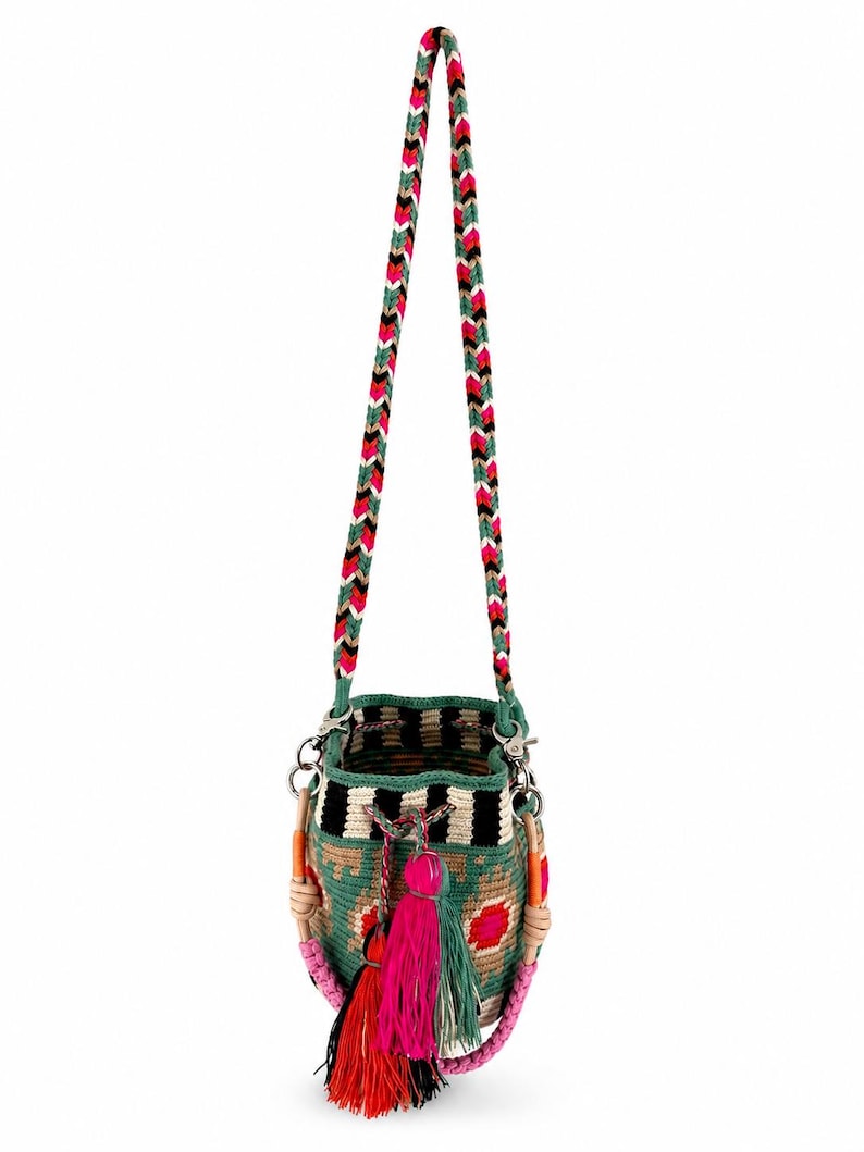 May include: A bucket bag featuring a woven green, black, and beige geometric pattern with a long braided strap in pink, green, white, and black. The bag includes a drawstring closure with multiple hanging tassels in red, pink, and green colors.