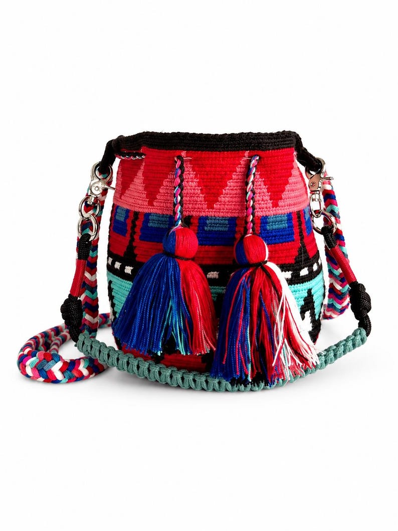 May include: A cylindrical crochet bucket bag featuring a vibrant geometric pattern in red, pink, blue, black, and white. The bag includes two large decorative tassels with matching colors and a thick, braided shoulder strap with silver hardware.