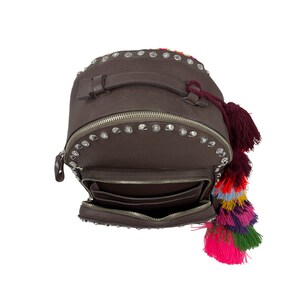 K&ouml;nnte beinhalten: Ein brauner Lederrucksack mit silbernen Nieten und bunten Quasten. Der Rucksack hat eine Rei&szlig;verschlusstasche vorne und einen Tragegriff oben.