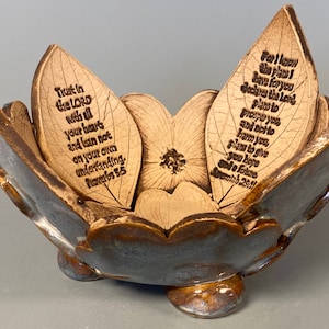 May include: A ceramic bowl with a floral design and three small feet. The bowl is brown and blue with a glossy finish. The interior features leaf-shaped elements with engraved Bible verses. The text includes "Trust in the Lord" and "For I know the plans I have for you."