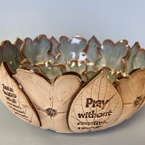 May include: A ceramic bowl with a floral design. The bowl is beige with a light blue interior and gold trim. The exterior features embossed floral petals with the text "Pray without ceasing."