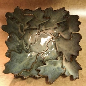 May include: A square ceramic bowl with a leaf design. The bowl is a muted green color with a glossy finish and brown trim. The leaves are layered to create a unique, textured appearance.