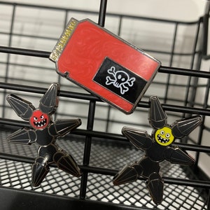 May include: Three enamel pins featuring a red and black can with a skull and crossbones, a black star-shaped robot with a red face, and a black star-shaped robot with a yellow face.