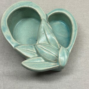 May include: A turquoise ceramic heart-shaped dish with two circular compartments and a stylized leaf design. The dish has a glossy finish and visible crackling throughout the surface, adding to its vintage aesthetic. The overall design is symmetrical and decorative.