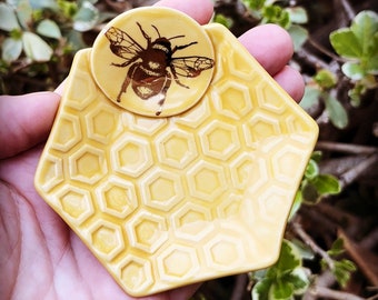 May include: A yellow ceramic trinket dish shaped like a hexagon with a raised honeycomb pattern across the surface. A small, circular, raised yellow accent piece is attached to the top edge, featuring a detailed dark brown illustration of a bee.