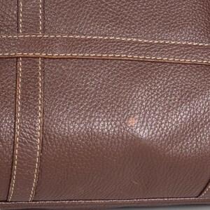 May include: Close-up of a brown leather bag with a textured surface and white stitching. The bag has a slight discoloration on the surface.
