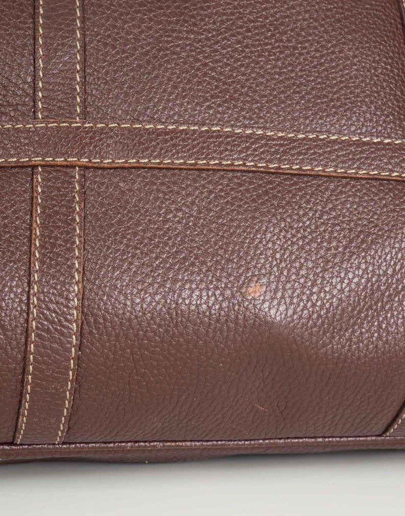May include: Close-up of a brown leather bag with a textured surface and white stitching. The bag has a slight discoloration on the surface.