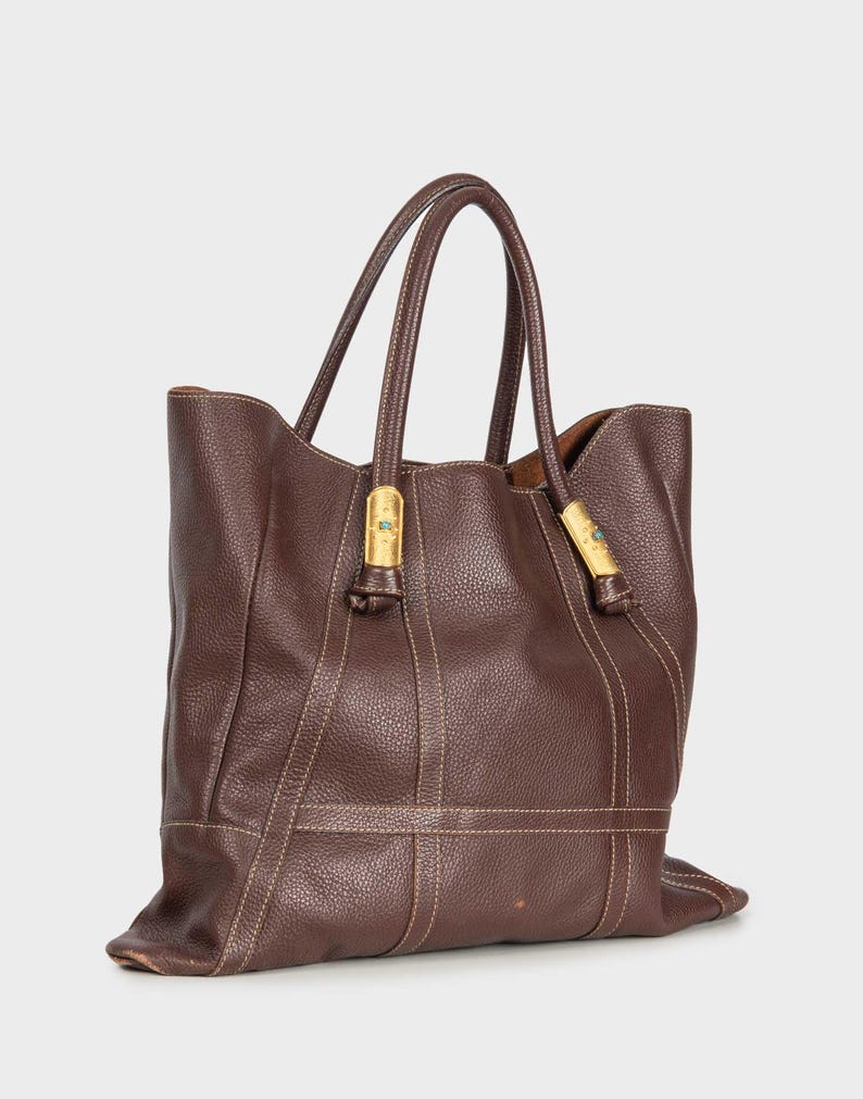 May include: A brown leather tote bag with gold hardware and a brown leather strap. The bag has a structured shape and a spacious interior.
