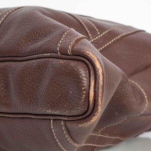 May include: Close-up of a brown leather handbag with white stitching. The bag has a textured surface and a slightly worn appearance.