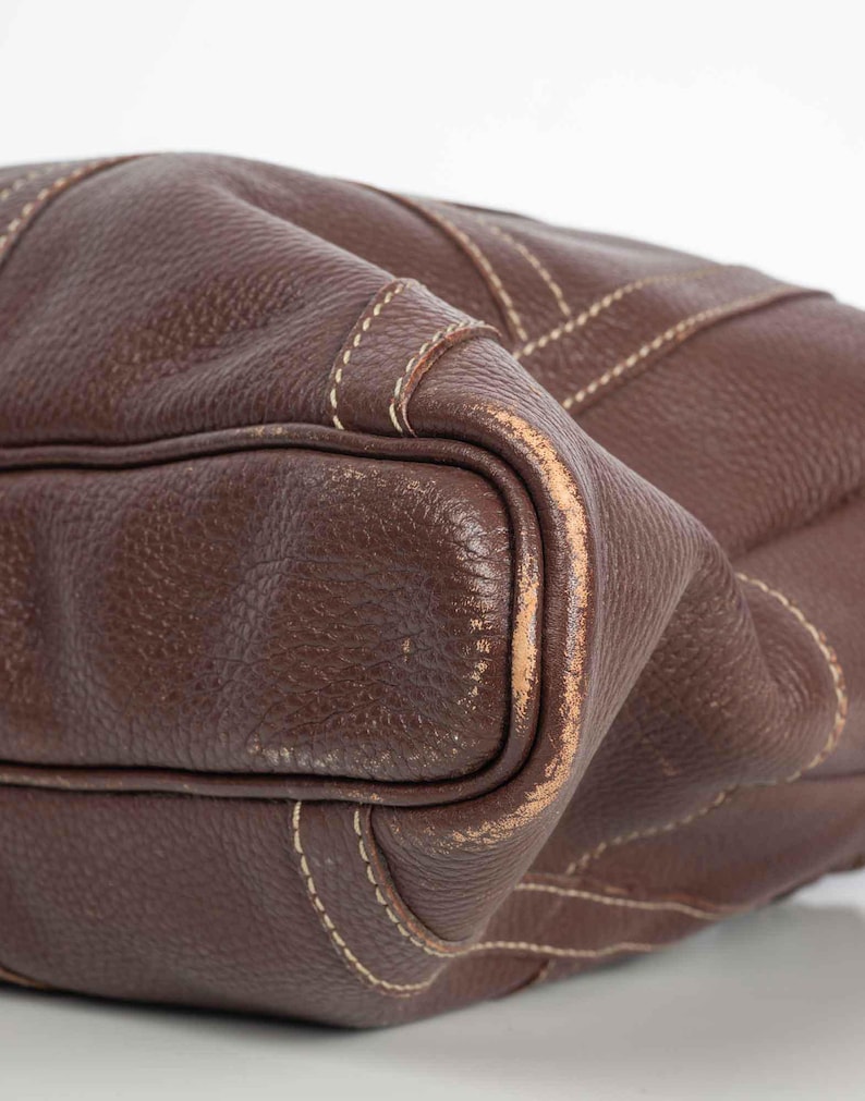 May include: Close-up of a brown leather handbag with white stitching. The bag has a textured surface and a slightly worn appearance.