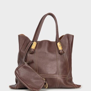 Una borsa tote di grandi dimensioni in pelle marrone scuro con una texture leggermente granulosa è presentata frontalmente. La borsa ha due manici superiori arrotondati fissati con dettagli in metallo dorato e elementi decorativi blu. Cuciture chiare a contrasto sono visibili lungo i bordi e in verticale sulla borsa. Una piccola pochette rettangolare in pelle marrone abbinata è agganciata. L'interno della borsa è di colore marrone chiaro.