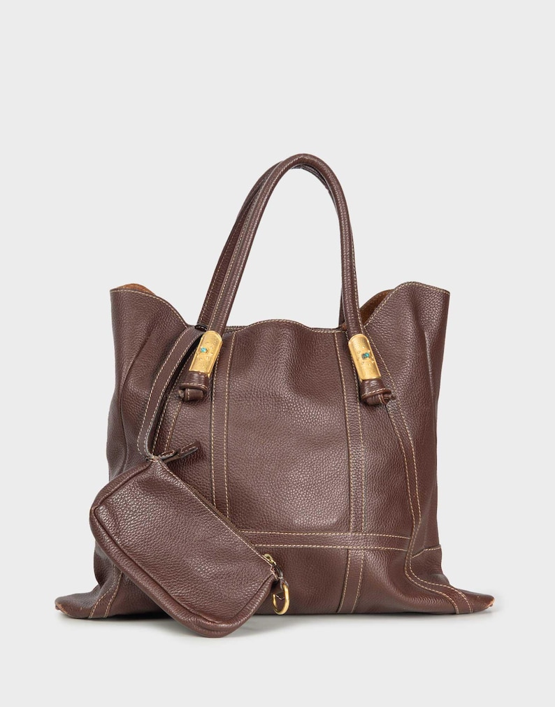 Una borsa tote di grandi dimensioni in pelle marrone scuro con una texture leggermente granulosa è presentata frontalmente. La borsa ha due manici superiori arrotondati fissati con dettagli in metallo dorato e elementi decorativi blu. Cuciture chiare a contrasto sono visibili lungo i bordi e in verticale sulla borsa. Una piccola pochette rettangolare in pelle marrone abbinata è agganciata. L'interno della borsa è di colore marrone chiaro.