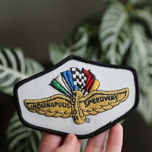 May include: Embroidered patch featuring the Indianapolis Motor Speedway logo. The patch has a white background with a black border. The logo includes gold wings, the words "Indianapolis Motor Speedway", and colorful flags.