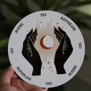 May include: A white, round divination board featuring a crescent moon and sunburst design. Two black hands are reaching towards the center. The words "Yes", "No", "Maybe", and "Rephrase" are printed around the edge.