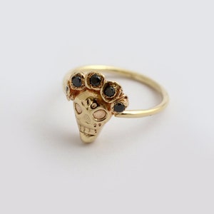 Puede incluir: Un anillo de oro con un dise&ntilde;o de calavera detallado y piedras preciosas negras alrededor de la parte superior.