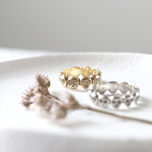 May include: Two skull rings, one gold and one silver, with small clear stones for eyes. The rings are on a white surface.