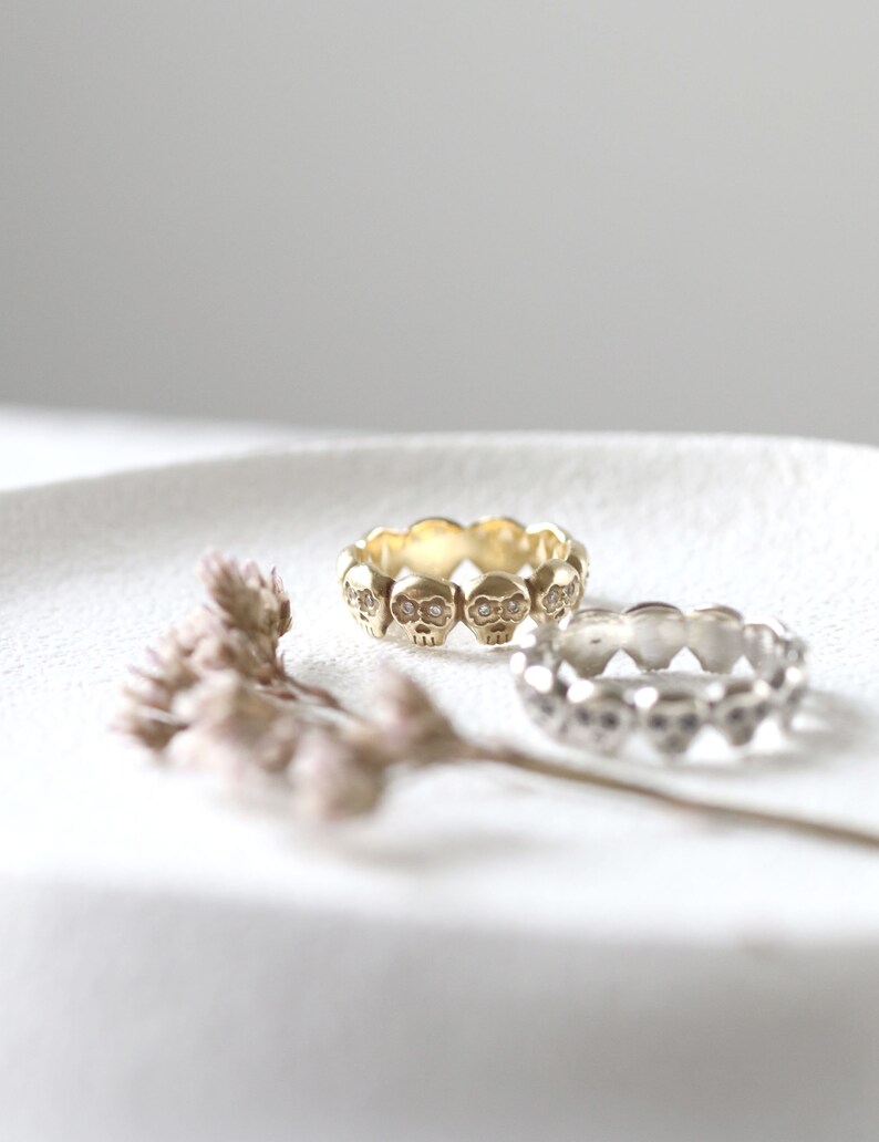 May include: Two skull rings, one gold and one silver, with small clear stones for eyes. The rings are on a white surface.
