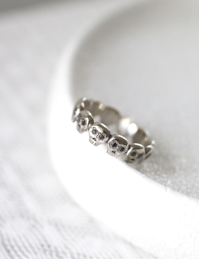 May include: A silver ring with a band of small skull designs, each skull has black diamond eyes.