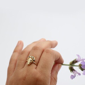 Puede incluir: Anillo de oro con un dise&ntilde;o triangular texturizado y piedras preciosas negras. El anillo se lleva en una mano que sostiene una flor morada.