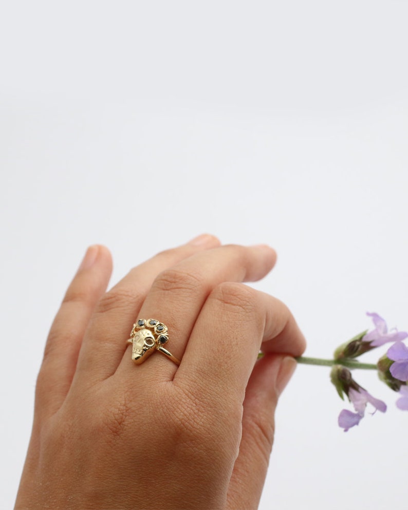 Puede incluir: Anillo de oro con un dise&ntilde;o triangular texturizado y piedras preciosas negras. El anillo se lleva en una mano que sostiene una flor morada.