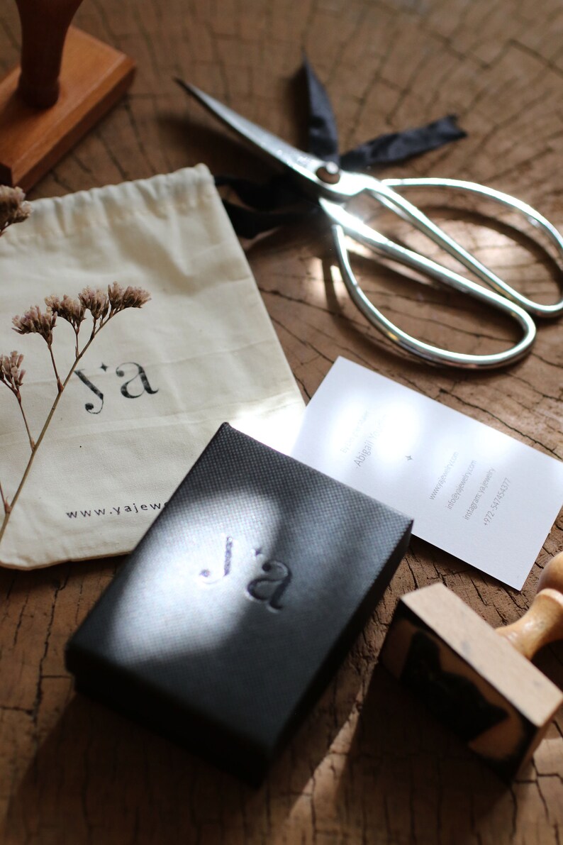 Puede incluir: Una caja negra con el texto "j'a" en relieve en la parte superior. La caja est&aacute; sobre una superficie de madera con una tarjeta blanca, una bolsa peque&ntilde;a y un sello de madera.