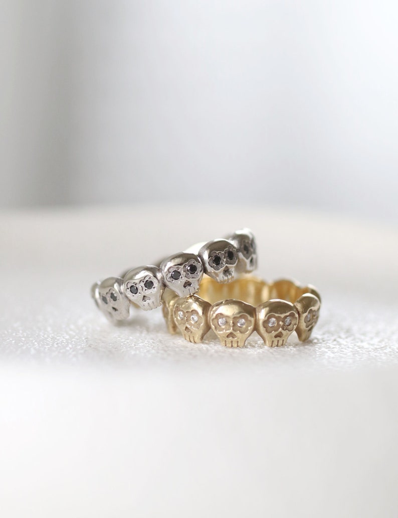 May include: Two skull rings with black and white diamonds. The top ring is silver with black diamonds and the bottom ring is gold with white diamonds.