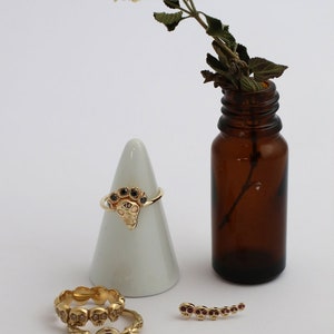 Puede incluir: Tres anillos de oro con dise&ntilde;os de calaveras y piedras preciosas negras y rojas. Los anillos se muestran en un cono blanco y una superficie blanca. Una peque&ntilde;a botella de vidrio marr&oacute;n con un tallo de flor seca est&aacute; en el fondo.