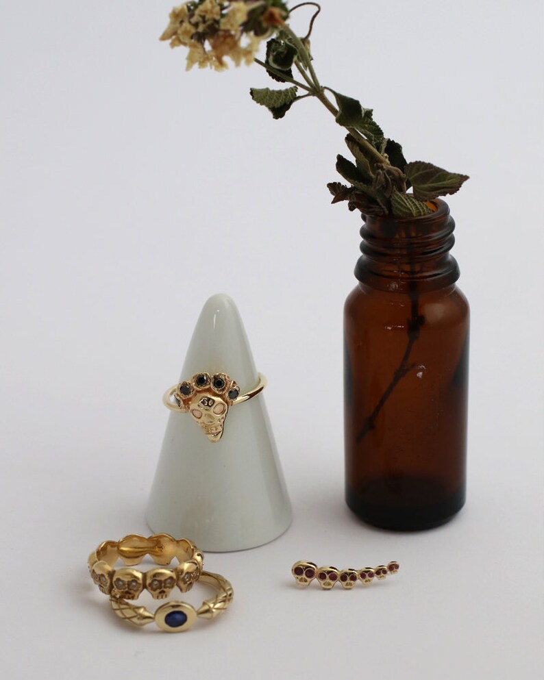 Puede incluir: Tres anillos de oro con dise&ntilde;os de calaveras y piedras preciosas negras y rojas. Los anillos se muestran en un cono blanco y una superficie blanca. Una peque&ntilde;a botella de vidrio marr&oacute;n con un tallo de flor seca est&aacute; en el fondo.