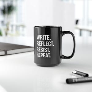 May include: A black ceramic mug with the words "WRITE. REFLECT. RESIST. REPEAT." in white lettering. The mug has a large handle and is sitting on a white surface.