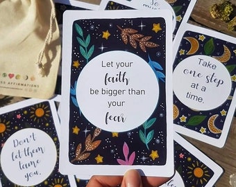 May include: A set of affirmation cards with a dark blue background and colorful illustrations of stars, moons, and leaves. The cards feature positive messages such as "Let your faith be bigger than your fear" and "Take one step at a time."