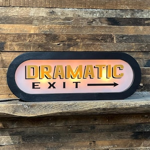 May include: An oval-shaped illuminated sign with a black frame and a white background. The text reads DRAMATIC in bold gold letters and EXIT in black letters, followed by a black arrow pointing to the right. The sign emits a warm glow.