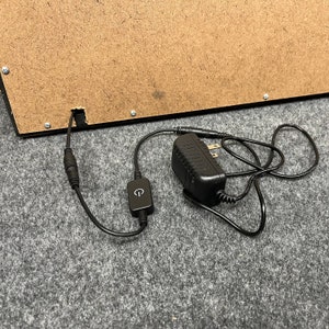 May include: Black power adapter with a black cord and a small black switch. The cord is plugged into a brown wooden box.