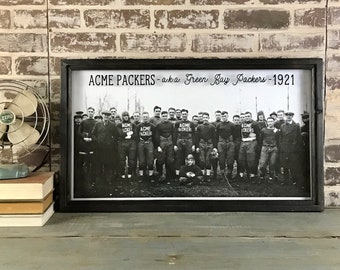May include: A black and white photo of the 1921 Acme Packers, also known as the Green Bay Packers, in a framed print. The text "ACME PACKERS - aka Green Bay Packers - 1921" is printed above the photo.