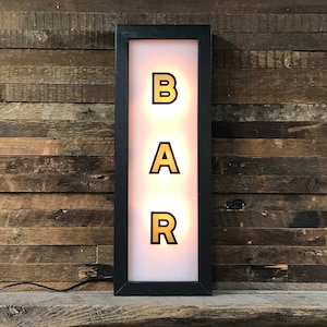 May include: A black framed sign with a white background and the word "BAR" in gold letters. The letters are illuminated from behind.