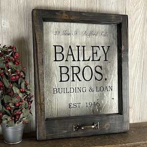 May include: A black framed sign with a white background and black text. The text reads "23 Main St. - Bedford Falls", "Bailey Bros.", "Building & Loan", and "Est. 1946".