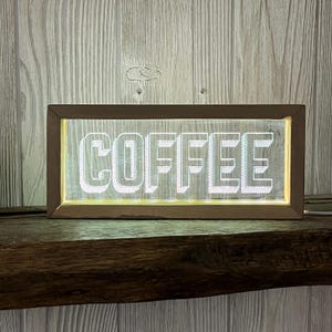 May include: A rectangular, illuminated sign with the word "COFFEE" in white, block letters. The sign has a brown wooden frame and is lit from within, creating a glowing effect. The sign is placed on a wooden shelf.