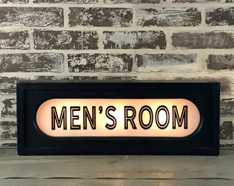 May include: Black rectangular sign with a white oval in the center. The oval has the text "MEN'S ROOM" in black lettering.