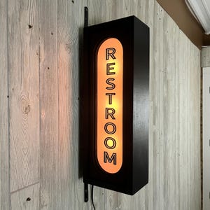 May include: A rectangular black wall-mounted sign with a vertical oval cutout in the center. The cutout features a glowing orange light behind a black frame with the word RESTROOM printed in bold black capital letters stacked vertically.