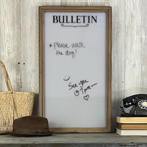 May include: A white dry erase board with a wooden frame. The board has the word "BULLETIN" written in black at the top. There are handwritten messages on the board, including "*Please walk the dog!" and "See you @ 1pm - &hearts;".