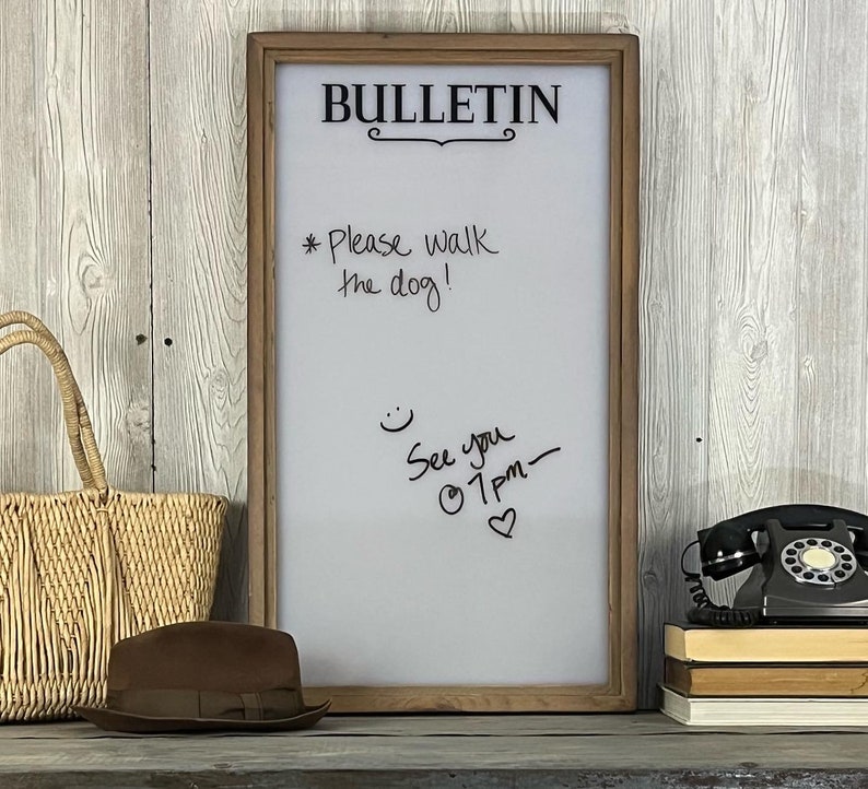May include: A white dry erase board with a wooden frame. The board has the word "BULLETIN" written in black at the top. There are handwritten messages on the board, including "*Please walk the dog!" and "See you @ 1pm - &hearts;".