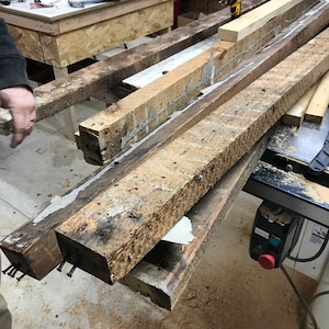 May include: Close-up of two weathered wooden beams joined together with glue. The beams are resting on a table saw with sawdust scattered around.