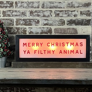 May include: A black framed light-up sign with the words "Merry Christmas Ya Filthy Animal" in red letters. The sign is sitting on a wooden shelf in front of a brick wall.