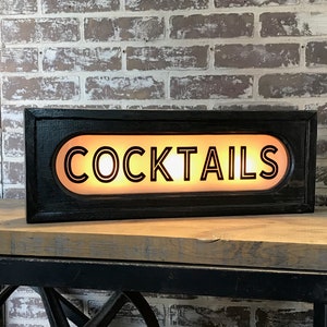 May include: Black wooden sign with a lit-up oval-shaped panel that reads "COCKTAILS" in white block letters with black outlines.