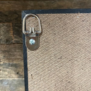 May include: A close-up of a silver hanging hook attached to the back of a wooden frame. The hook is designed to hang the frame on a wall.