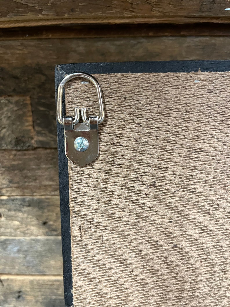 May include: A close-up of a silver hanging hook attached to the back of a wooden frame. The hook is designed to hang the frame on a wall.