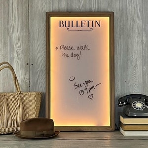 May include: A wooden framed dry erase board with a white surface and the word "BULLETIN" written in black at the top. The board has a warm yellow glow and is lit from behind. The board has handwritten messages in black marker, including "Please walk the dog!" and "See you @ 1pm."
