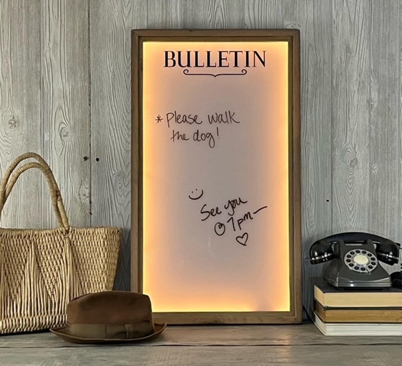 May include: A wooden framed dry erase board with a white surface and the word "BULLETIN" written in black at the top. The board has a warm yellow glow and is lit from behind. The board has handwritten messages in black marker, including "Please walk the dog!" and "See you @ 1pm."